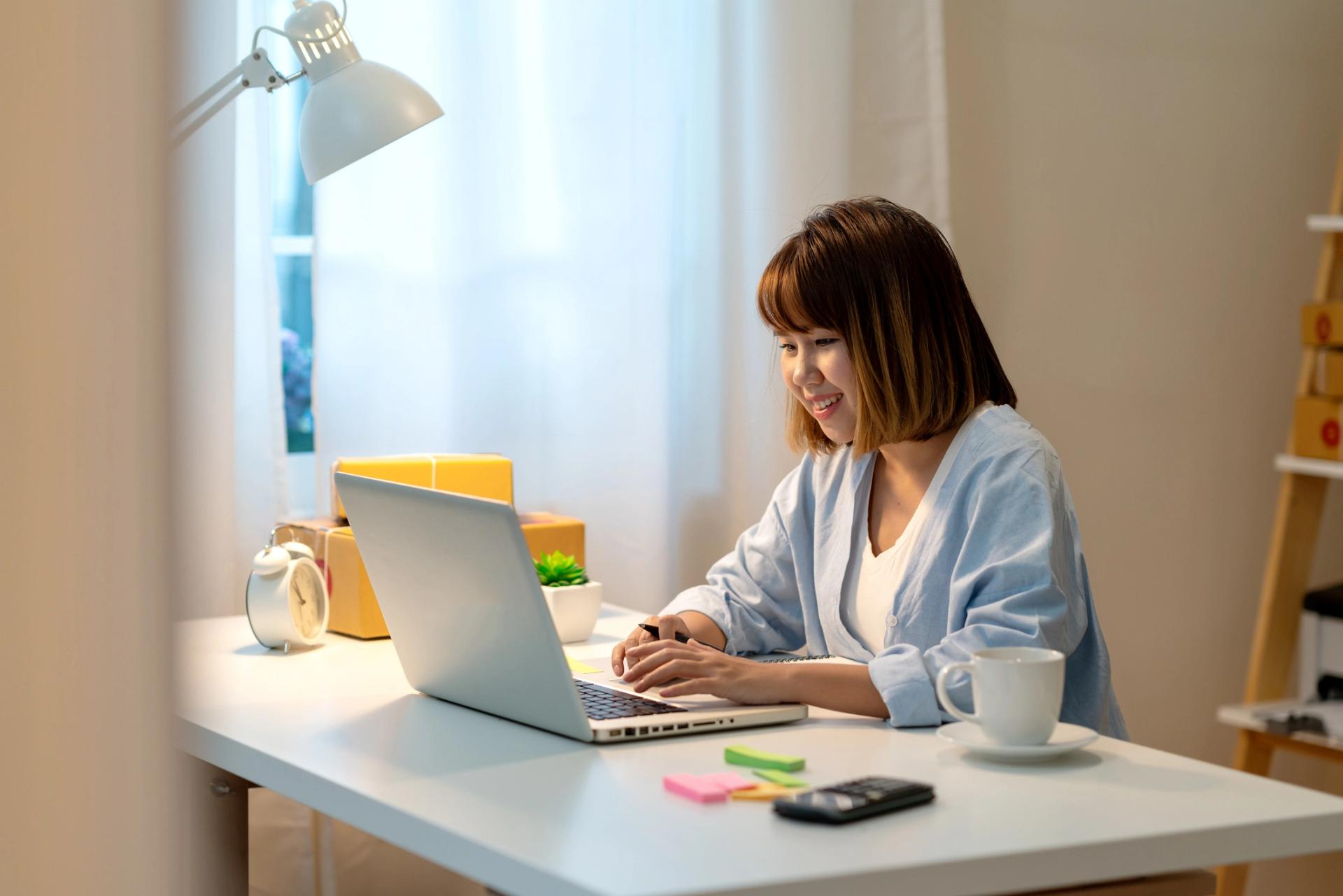 A woman smiling and working on her laptop at a desk in a cozy home environment.