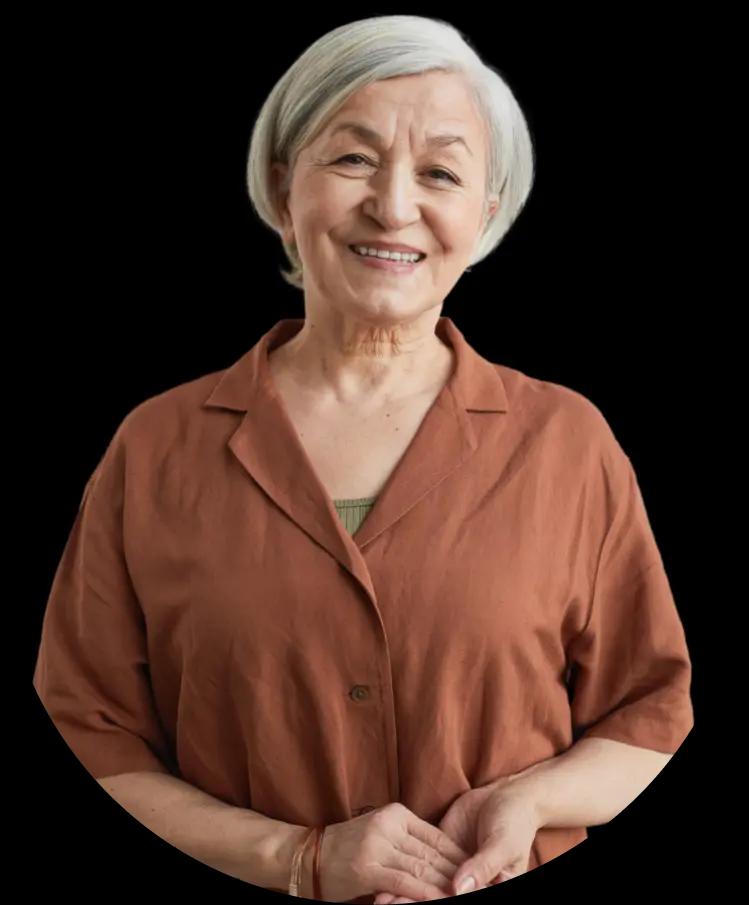 An elderly woman with light skin and short gray hair, wearing a brown blouse. She is smiling warmly.