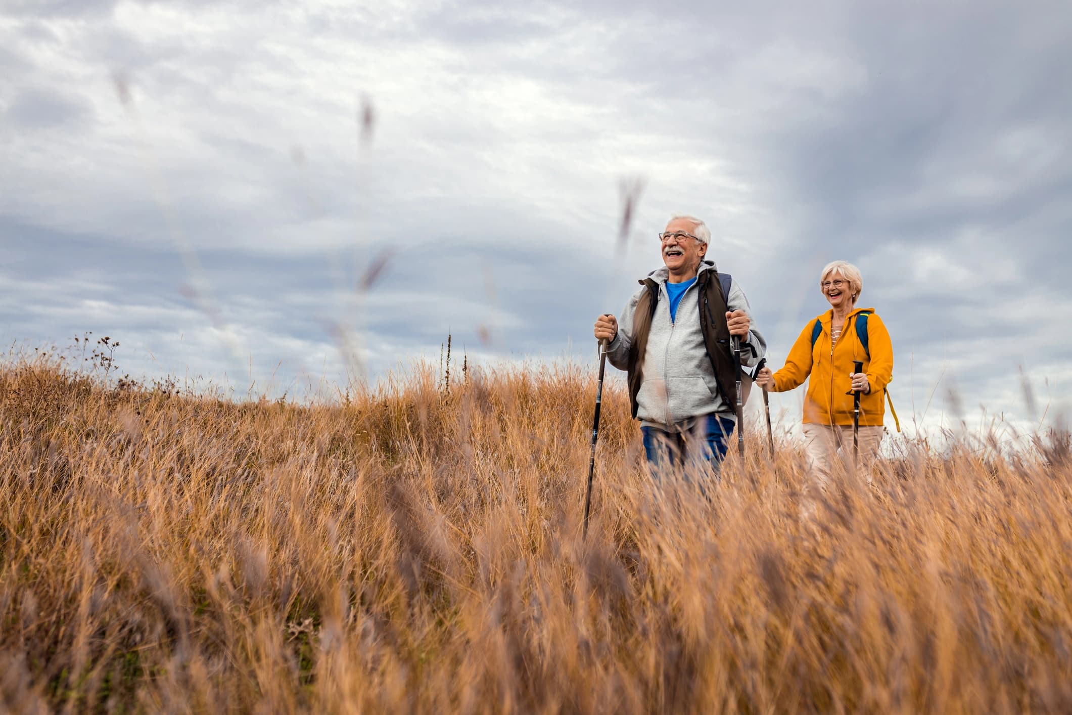 An elderly couple is hiking through a field of tall, dry grass under an overcast sky.