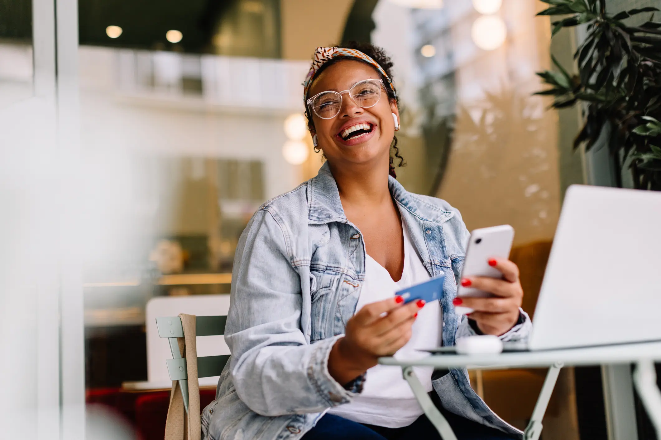 A joyful woman sitting at an outdoor café, wearing a light denim jacket and a headband.